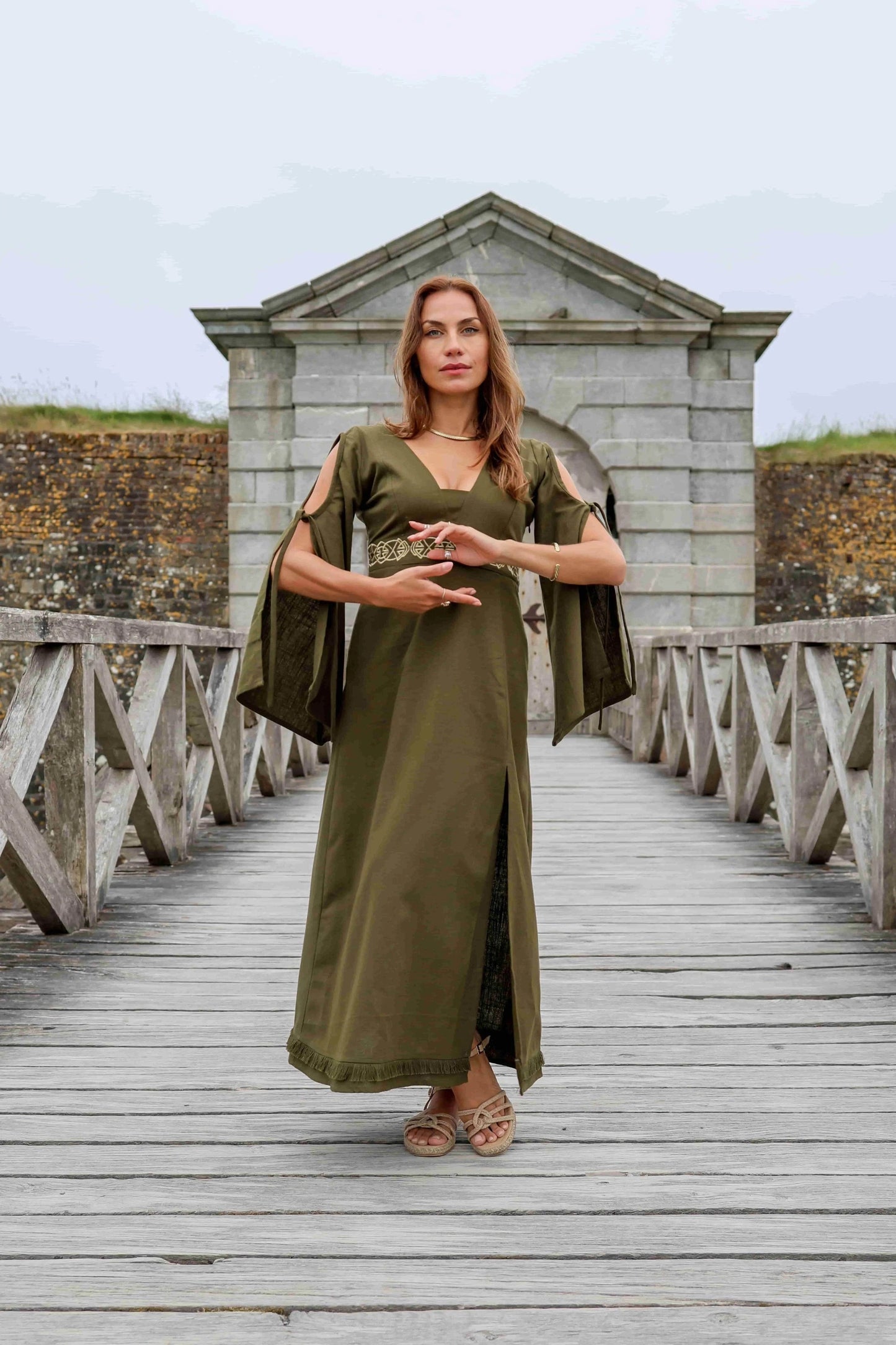Maeve Celtic Dress worn on wooden bridge, green Celtic maxi dress with bell sleeve detail and embroidered waist for formal folklore styling.
