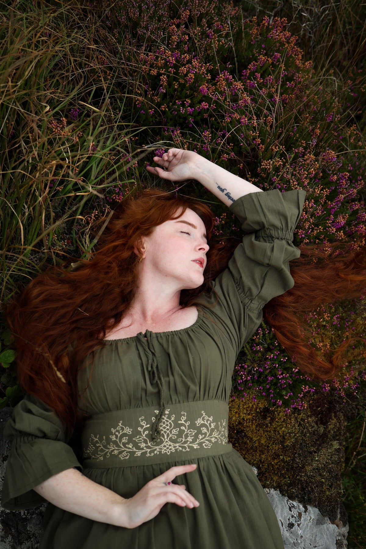 A woman with long red hair lies on her back in tall grass and among pink flowers, wearing a romantic sage green dress with a floral embroidered waistband and bunched sleeves