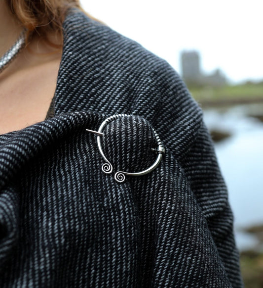 Close-up of handwoven shawl fastened with white metal Uisneach brooch, showing woven texture and silver spiral pin detail