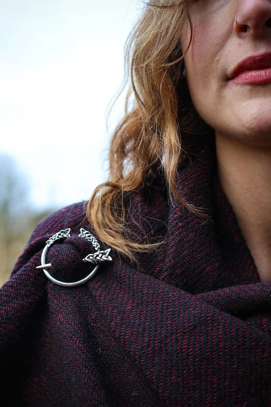 Close-up of maroon and black woven shawl with silver Trinity brooch showing Celtic knot detail and wool fabric texture