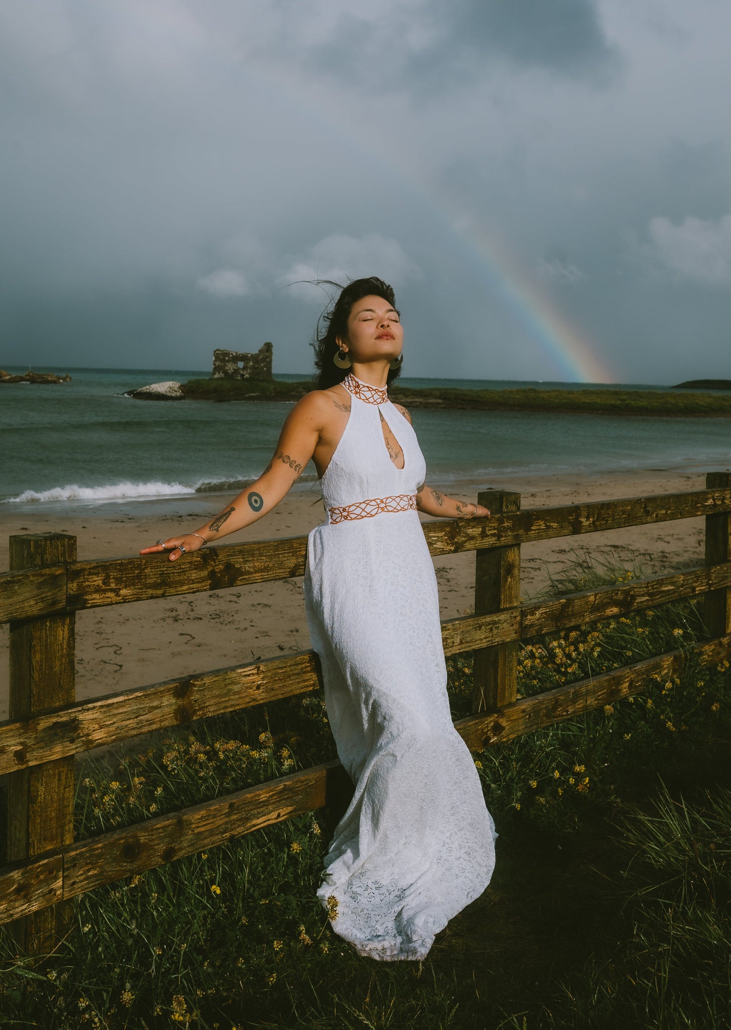 A woman in a flowing white halter maxi wedding dress with Celtic embroidery stands outdoors by a wooden fence near a beach, looking upward under a dramatic cloudy sky with a faint rainbow in the distance.