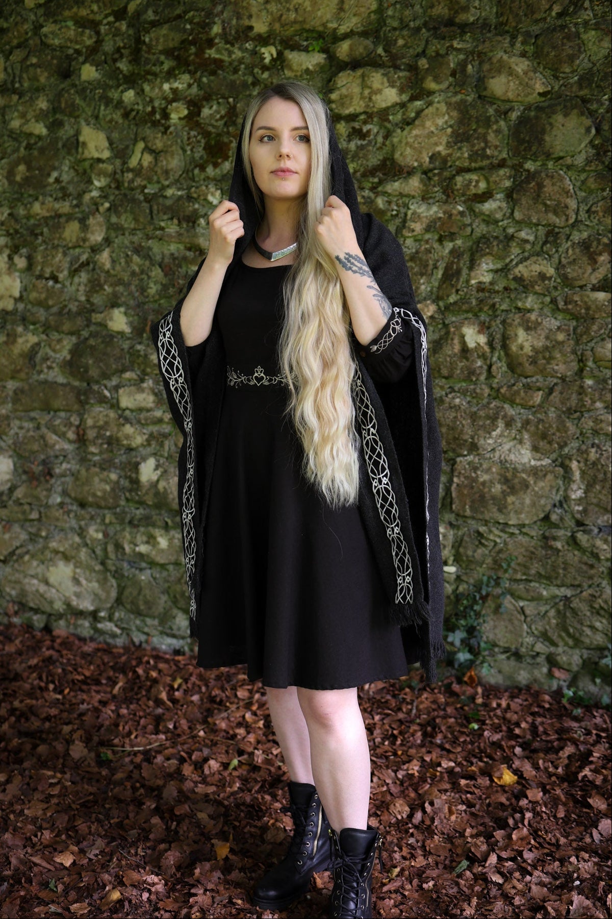 A woman with long blonde hair wearing a black medieval-style dress with a matching black hood cape with silver embroidered trim, standing in front of a rustic stone wall on a bed of brown leaves.