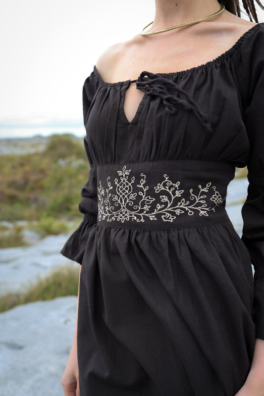 Black cotton folklore dress showing delicate botanical and Celtic knotwork embroidery across the fitted waist panel