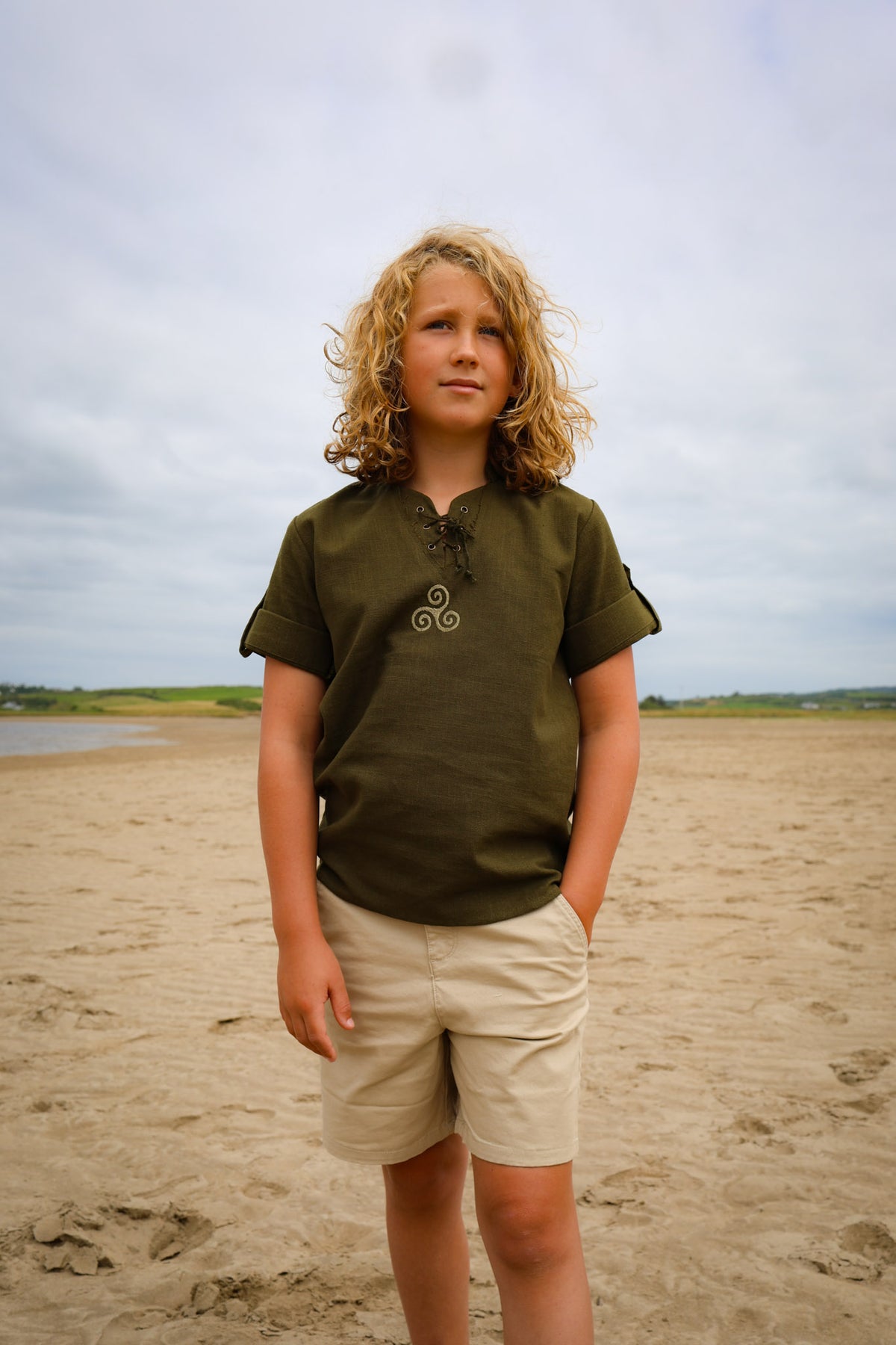 Boys Celtic Medieval Shirt in olive green with Triskele embroidery, lace-up neck and roll-up sleeves, styled outdoors on an Irish beach