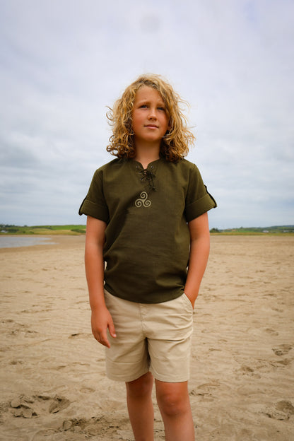 Boys Celtic Medieval Shirt in olive green with Triskele embroidery, lace-up neck and roll-up sleeves, styled outdoors on an Irish beach