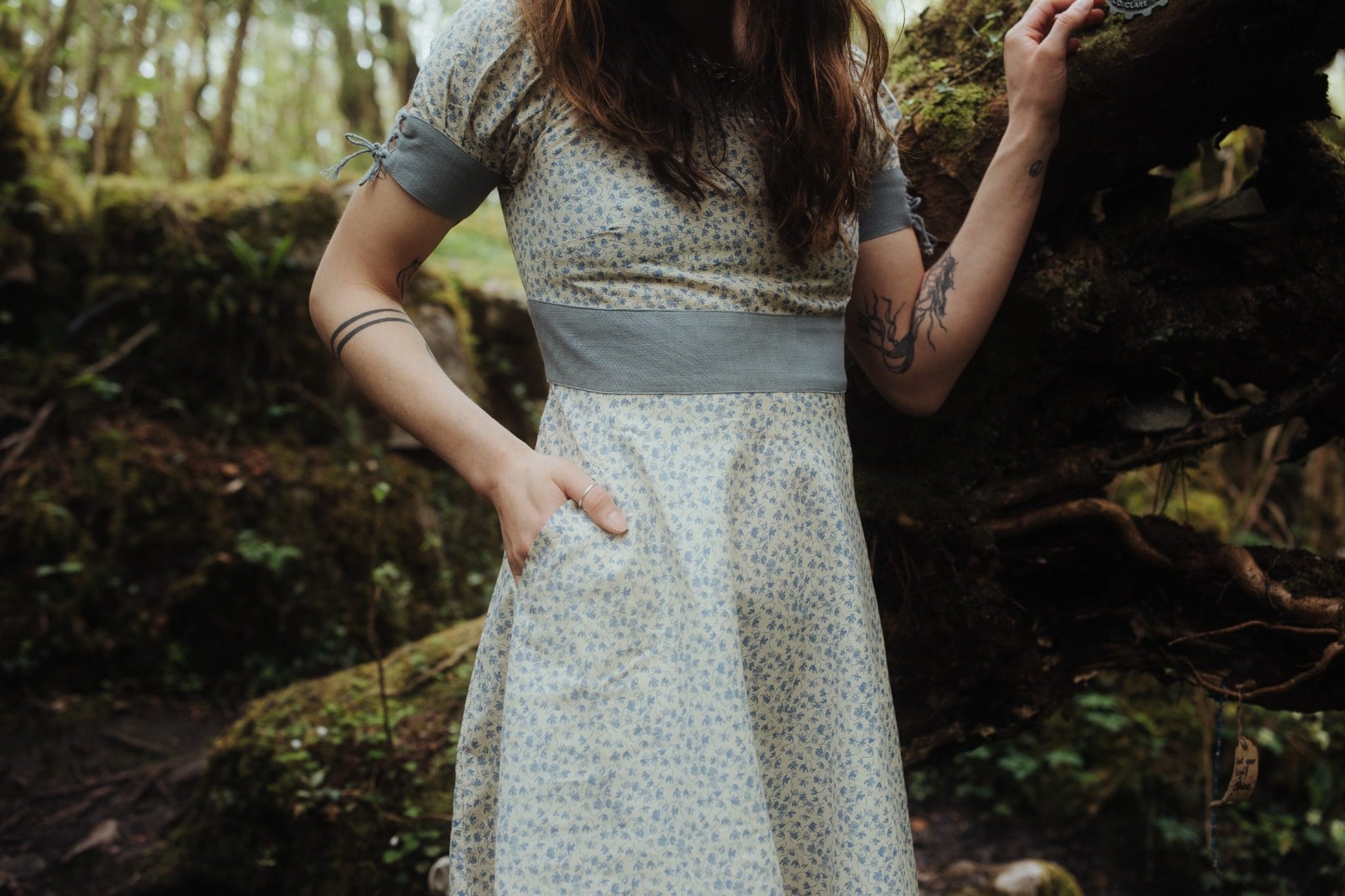 Forget Me Not Linen Dress with pockets shown in a woodland setting, featuring blue floral print fabric, fitted waist, corset back and soft folklore-inspired styling.