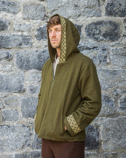 Man wearing a green hooded jacket with celtic embroidered details against a stone wall.