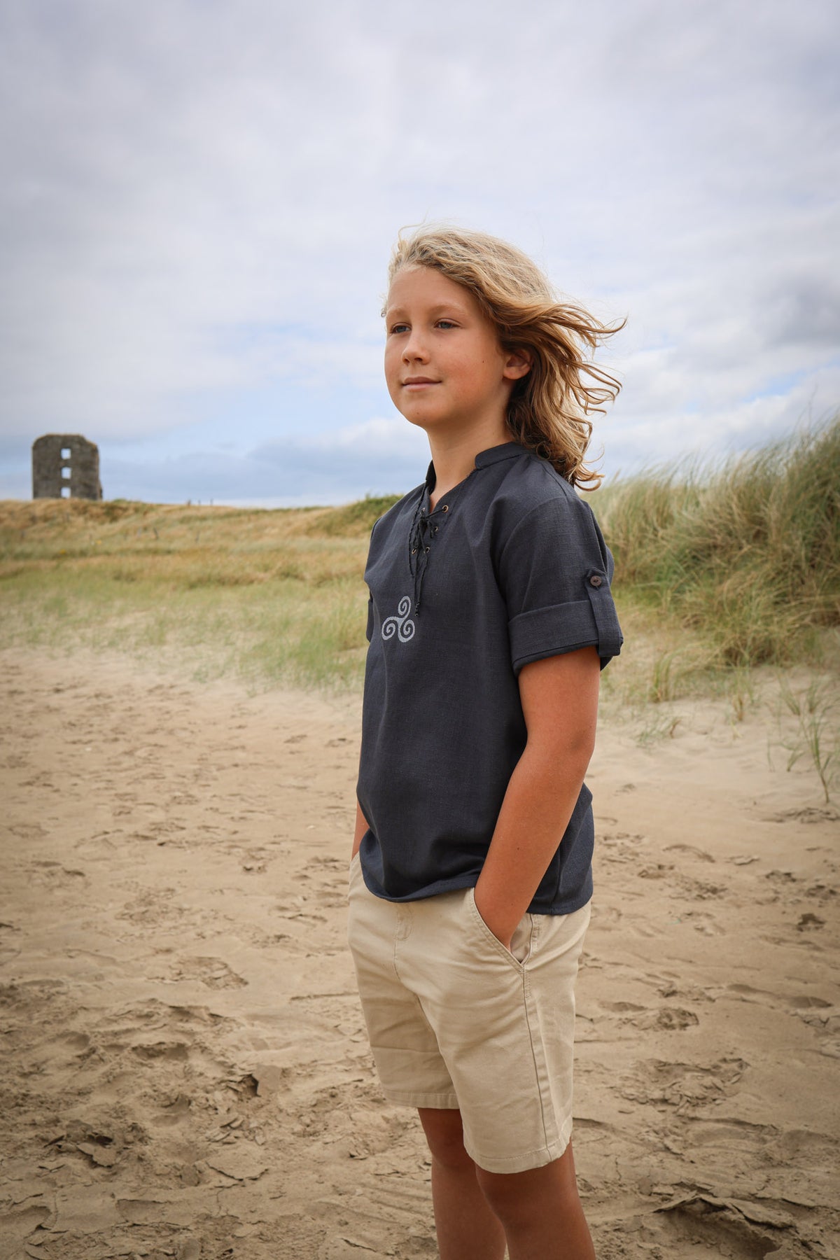 Navy blue boys Celtic Triskele shirt with lace-up neckline and buttoned roll-up sleeves, styled on an Irish beach with Celtic Fusion clothing