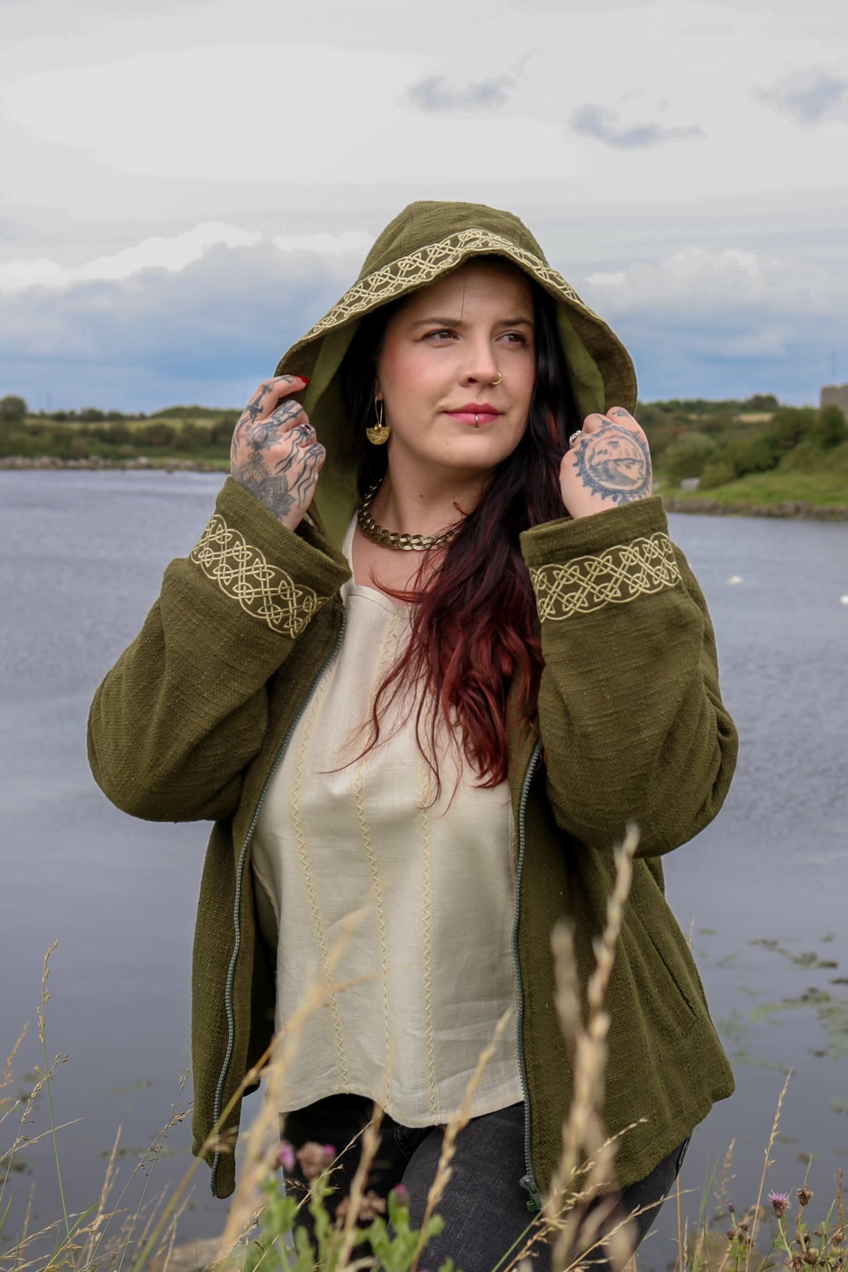 Olive green folklore jacket with deep hood, front zip and golden Celtic knotwork embroidery worn beside water.