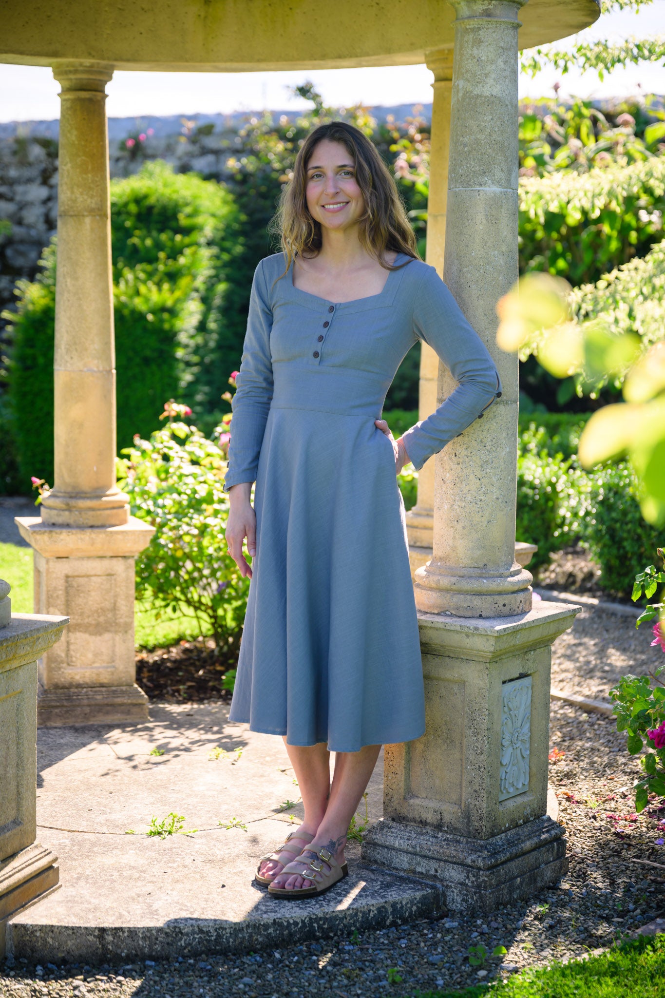 Pastel blue corset midi dress with pockets, long sleeves and a flowing circle skirt in a sunny garden.