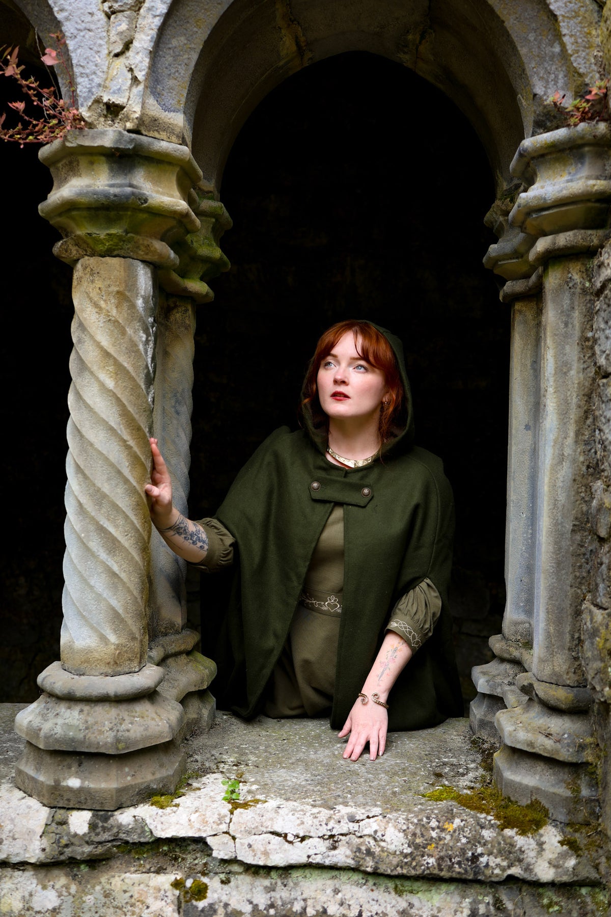 Irish Woman wearing green folklore wool cloak with hood and sleeveless poncho-style design in an old stone arch way window