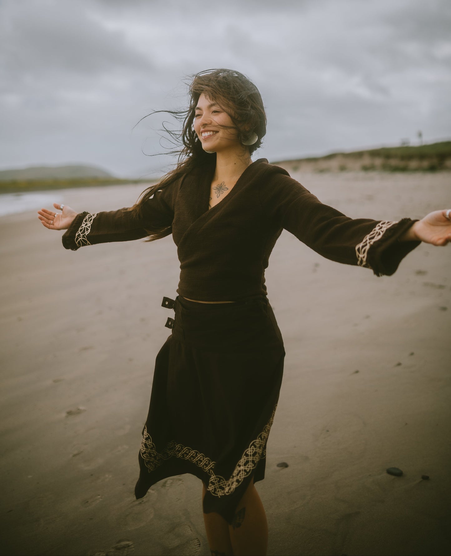 Brown folklore crop top with adjustable wrap fit and Celtic embroidered cuffs designed by Celtic Fusion and styled on a moody coastal beach with windswept hair