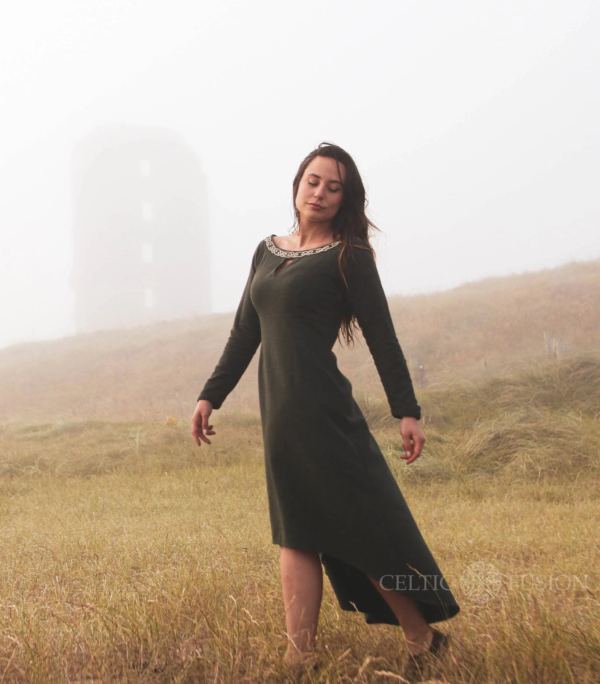 Green Celtic dress with Celtic knot embroidery, worn in an Irish landscape as an alternative wedding guest dress with high low skirt.