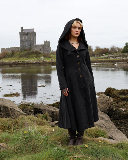 Woman wearing black Princess heavy wool maxi coat with oversized hood, button front and full flowing skirt beside an Irish castle