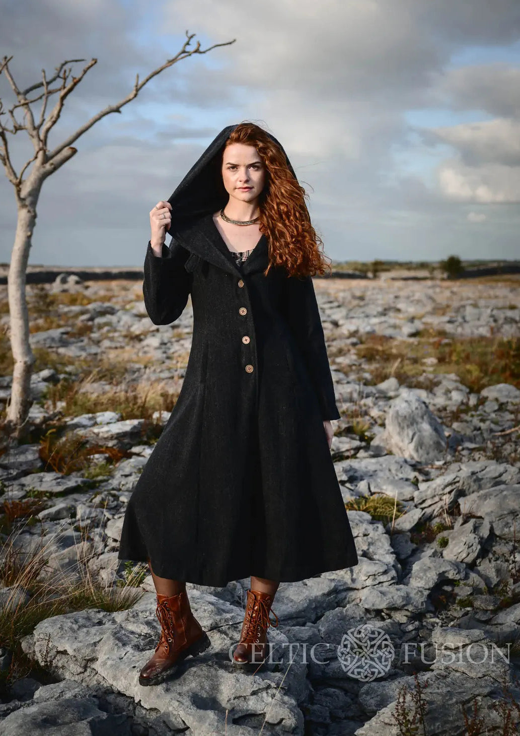Woman wearing black full-length hooded wool coat with fitted waist and flowing skirt on rocky Irish landscape in Ireland with Celtic Fusion