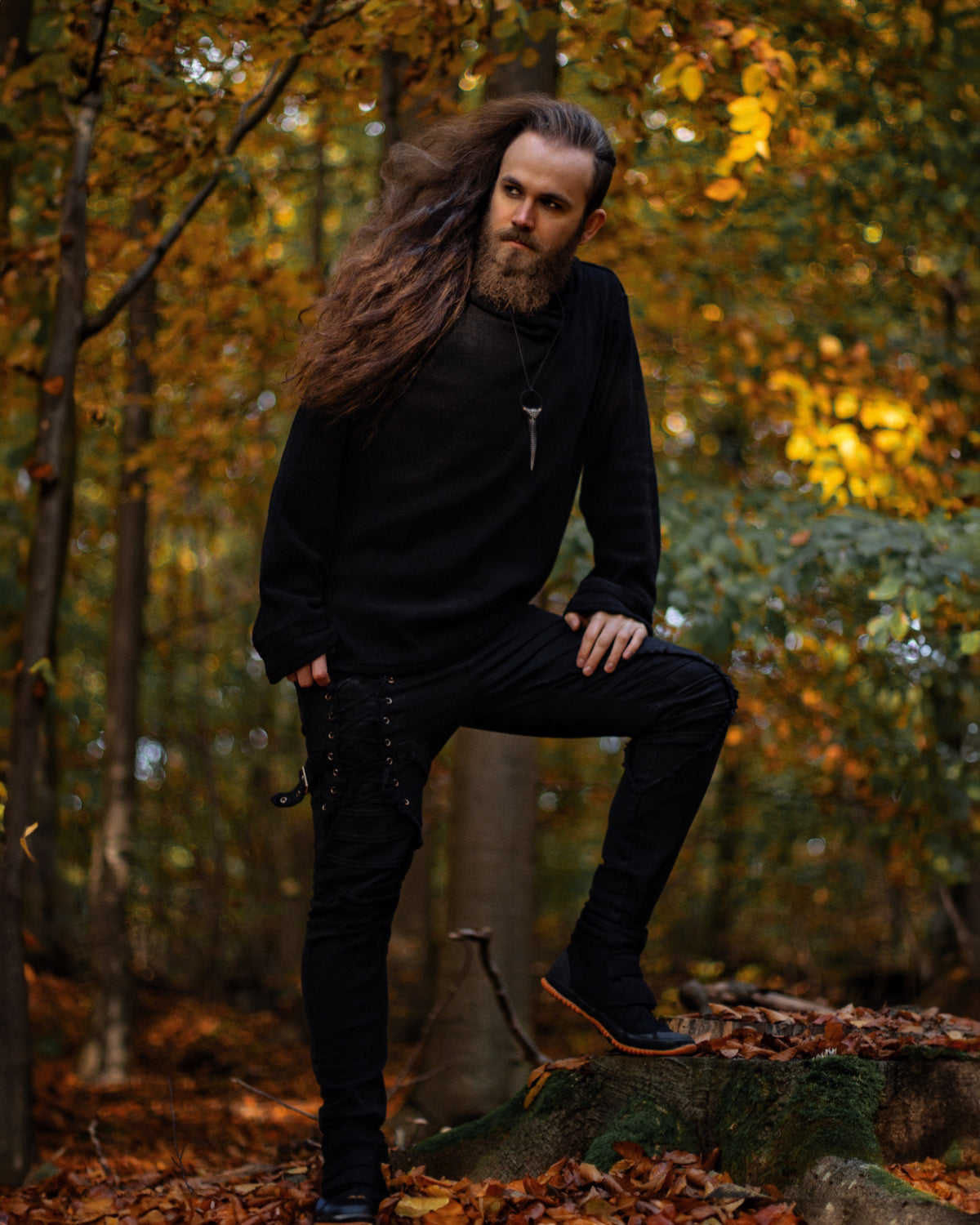 A man with long brown hair and a beard wearing a black long-sleeve hoodie, black pants, and black boots, posing on a tree stump in an autumn forest.
