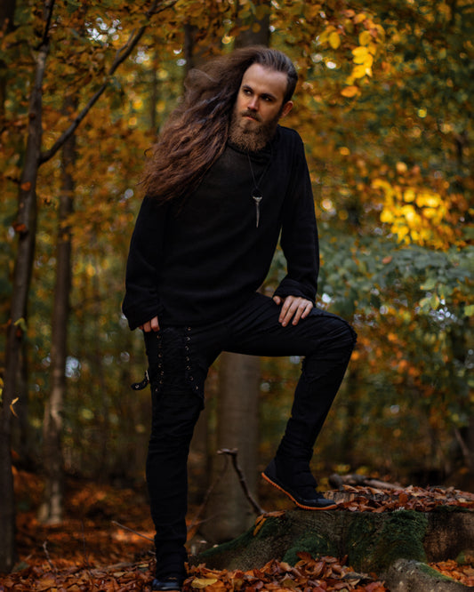 A man with long brown hair and a beard wearing a black long-sleeve hoodie, black pants, and black boots, posing on a tree stump in an autumn forest.