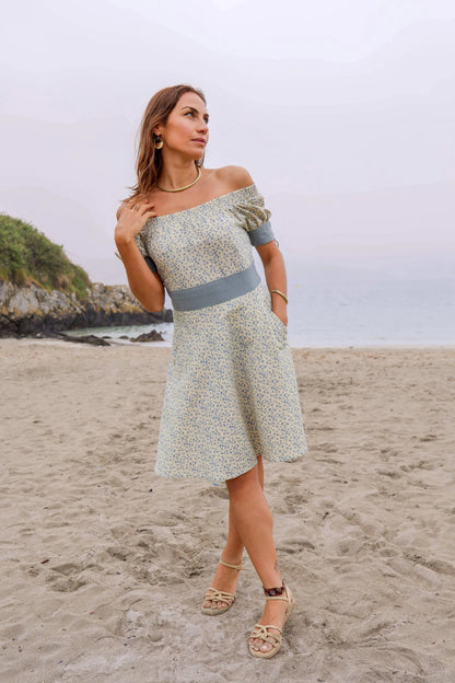 Forget Me Not Linen Dress styled on the beach, a blue floral linen blend dress with off-shoulder neckline, slate blue waistband and hidden pockets.