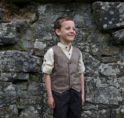 Boy wearing brown Tree of Life tweed waistcoat with cream shirt and dark trousers against a stone wall