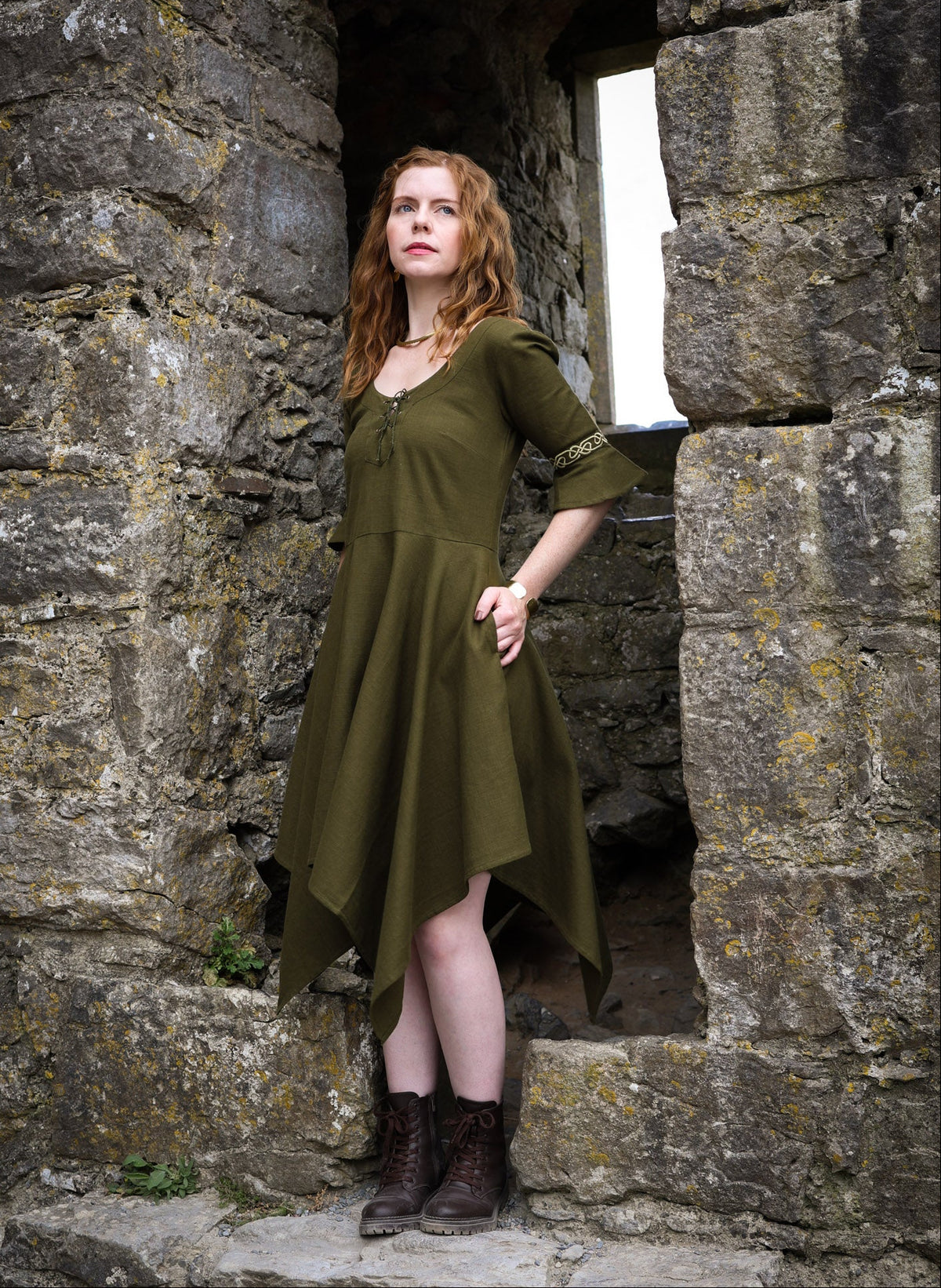 Green Macha corset back dress with Celtic embroidered sleeves and handkerchief hem, photographed beside old stone ruins.