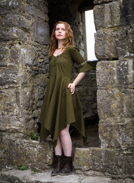 Green Macha corset back dress with Celtic embroidered sleeves and handkerchief hem, photographed beside old stone ruins.