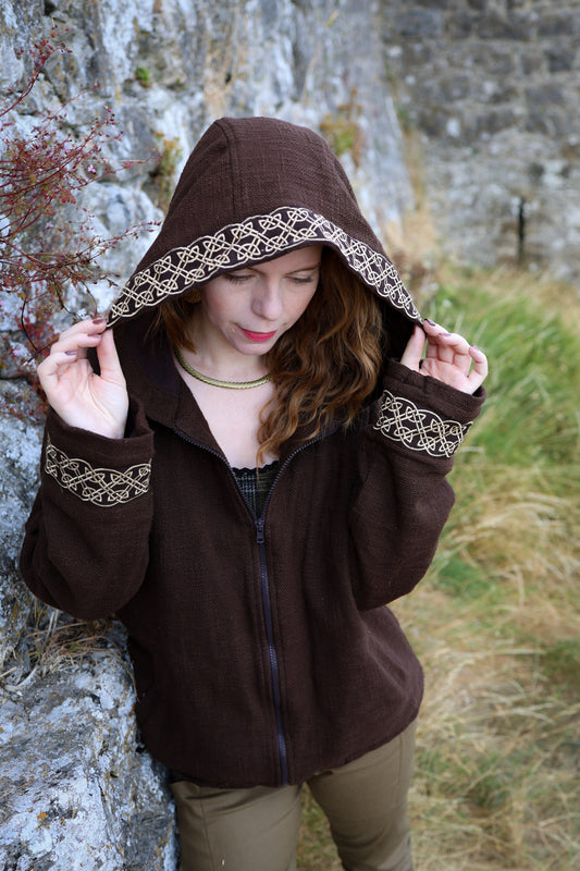 Close-up of brown Celtic hooded jacket showing embroidered knotwork trim around the deep hood and cuffs.
