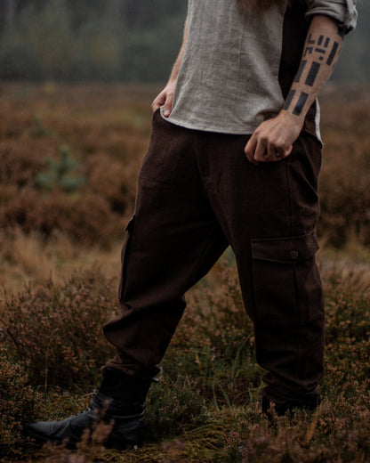 A person standing outdoors in a moody, grassy field wearing dark brown cargo pants, a light gray linen shirt, and black boots.