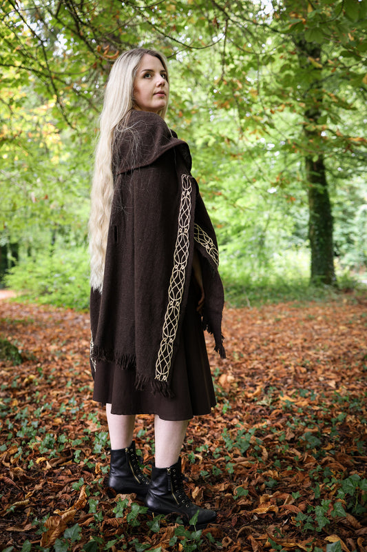 A woman with long blonde hair stands in a wooded outdoor setting, wearing a dark brown medieval-style cloak with gold geometric embroidery and black lace-up boots.