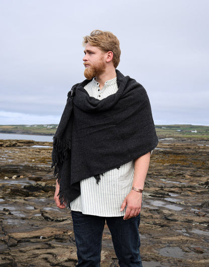 A man with a beard stands on a rocky coastal shoreline under an overcast sky, wearing a dark charcoal knit shawl draped over his shoulders, a white and black striped shirt, and dark jeans.