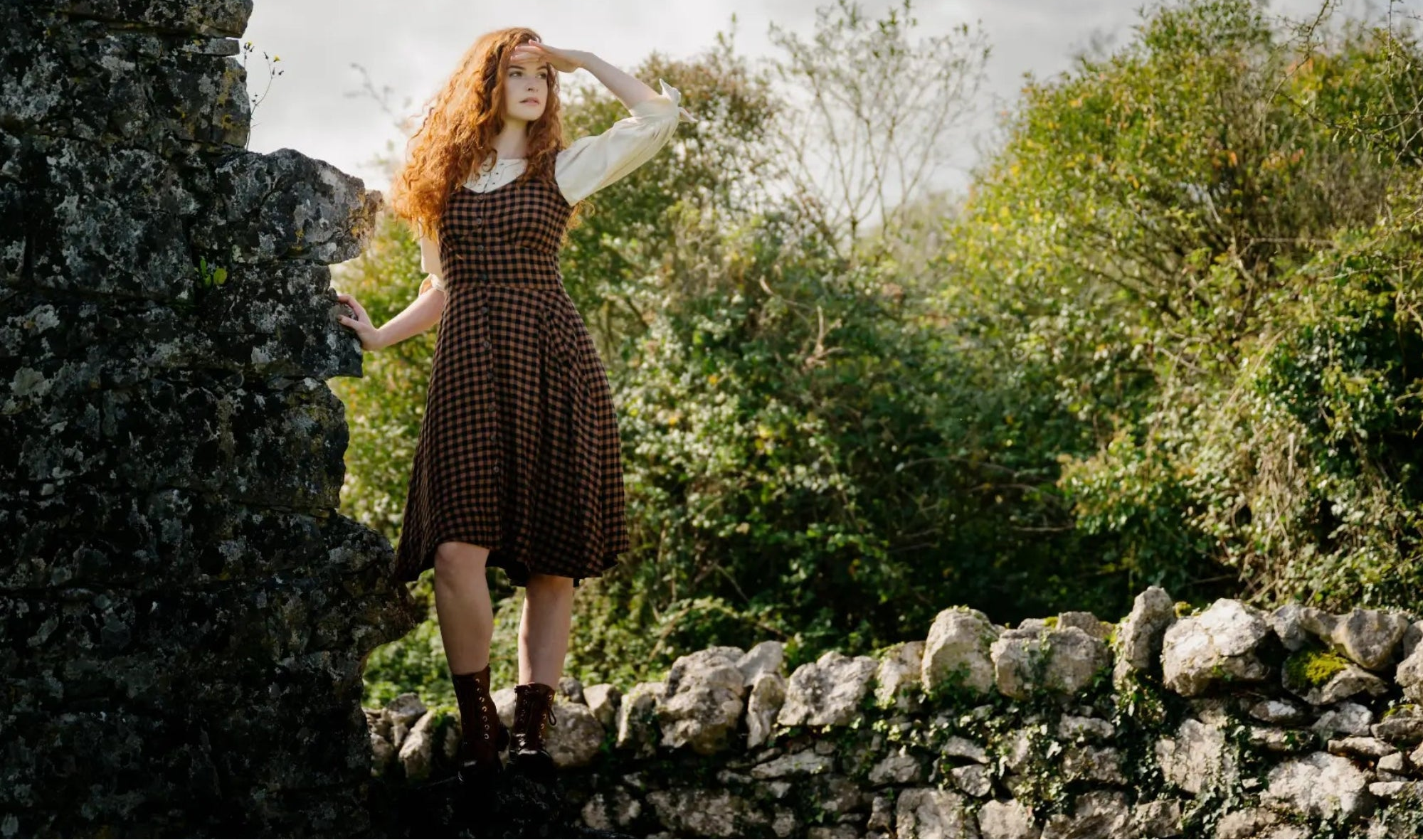 Celtic Fusion Design folklore inspired clothing, Irish designed women’s cottage core dress in gingham check styled beside old stone ruins in Ireland