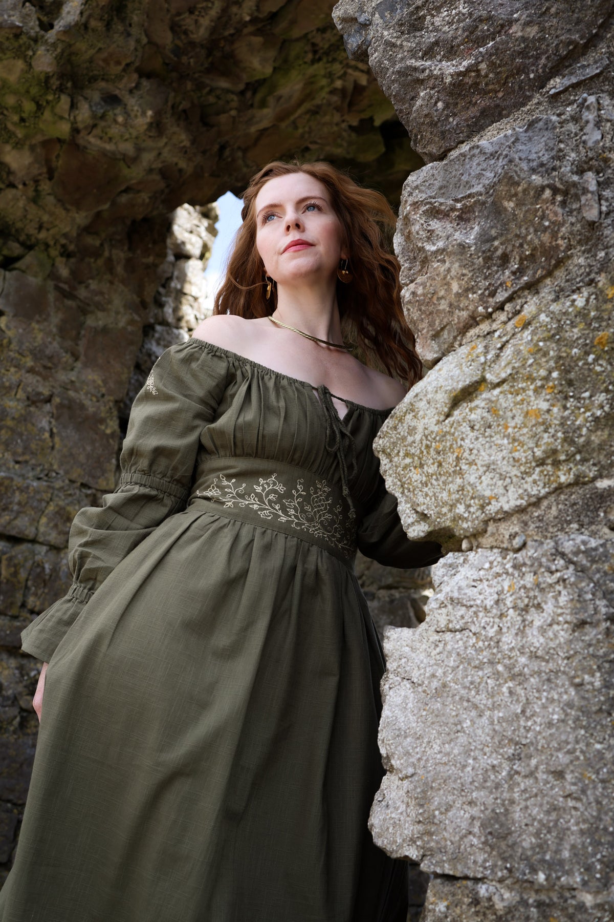 Sage green folklore midi dress with botanical embroidery, gathered bodice and long puff sleeves styled beside stone ruins.