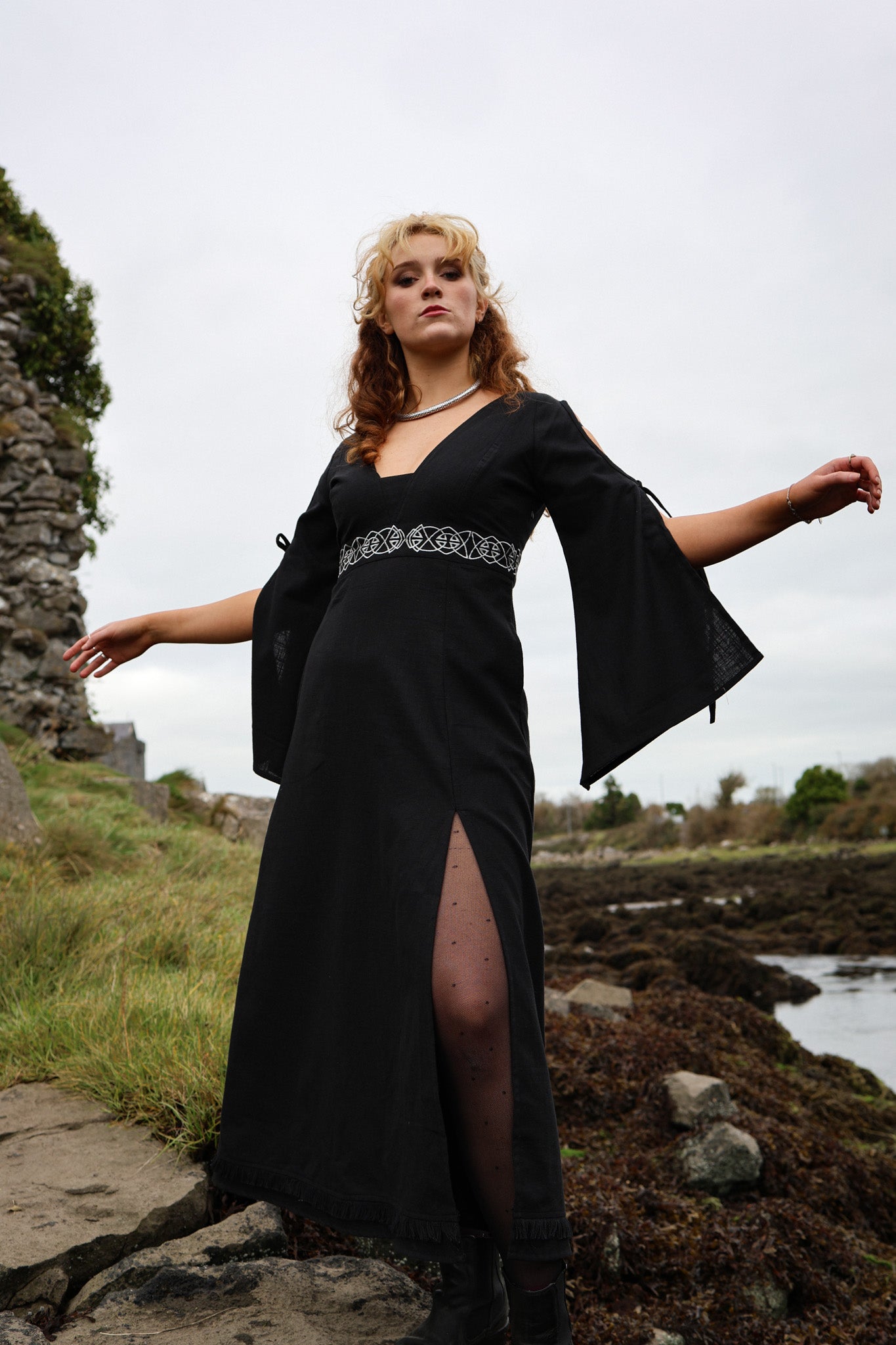 Maeve Black Celtic Dress in a coastal setting, front view of a gothic folklore dress with bell sleeves, Celtic knot embroidery and flowing black skirt