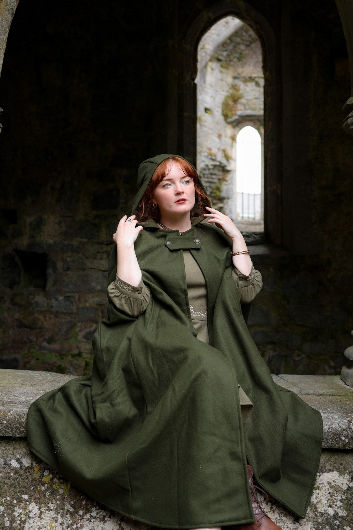 Woman wearing green folklore hooded wool cloak with long maxi length, pockets and sleeveless cape design outdoors