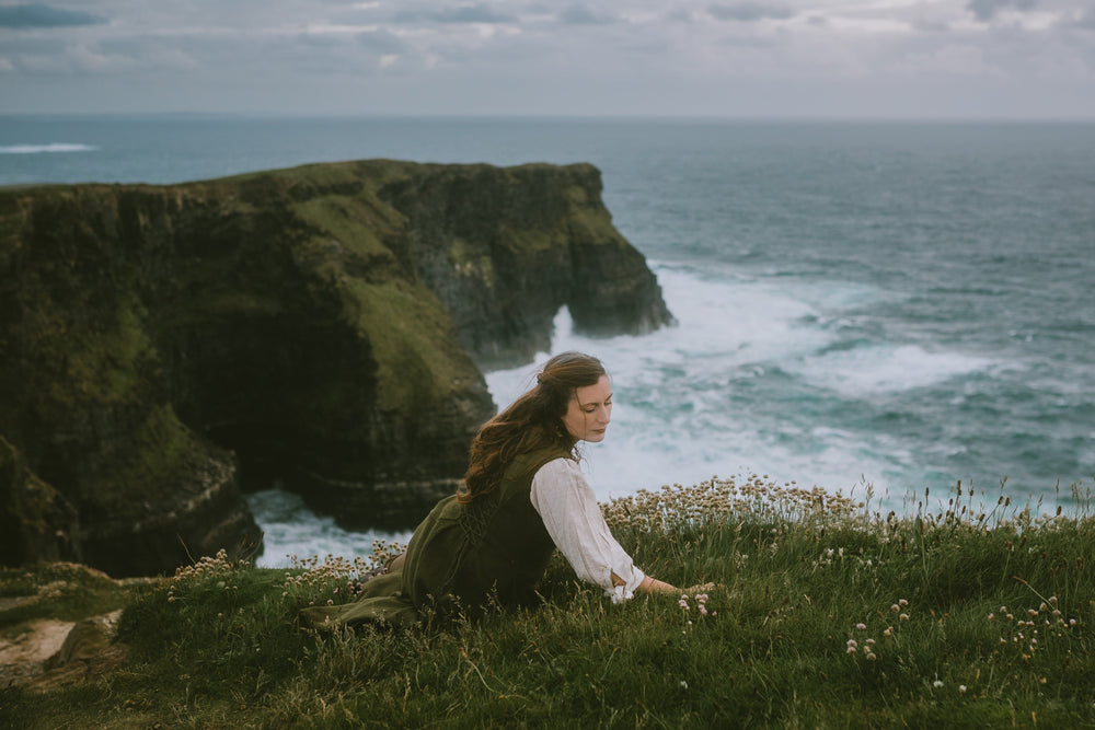 A woman in a natural cotton blouse and dark green vest dress sits on a grassy cliffside overlooking the cliffs of moher and ocean under a cloudy sky.