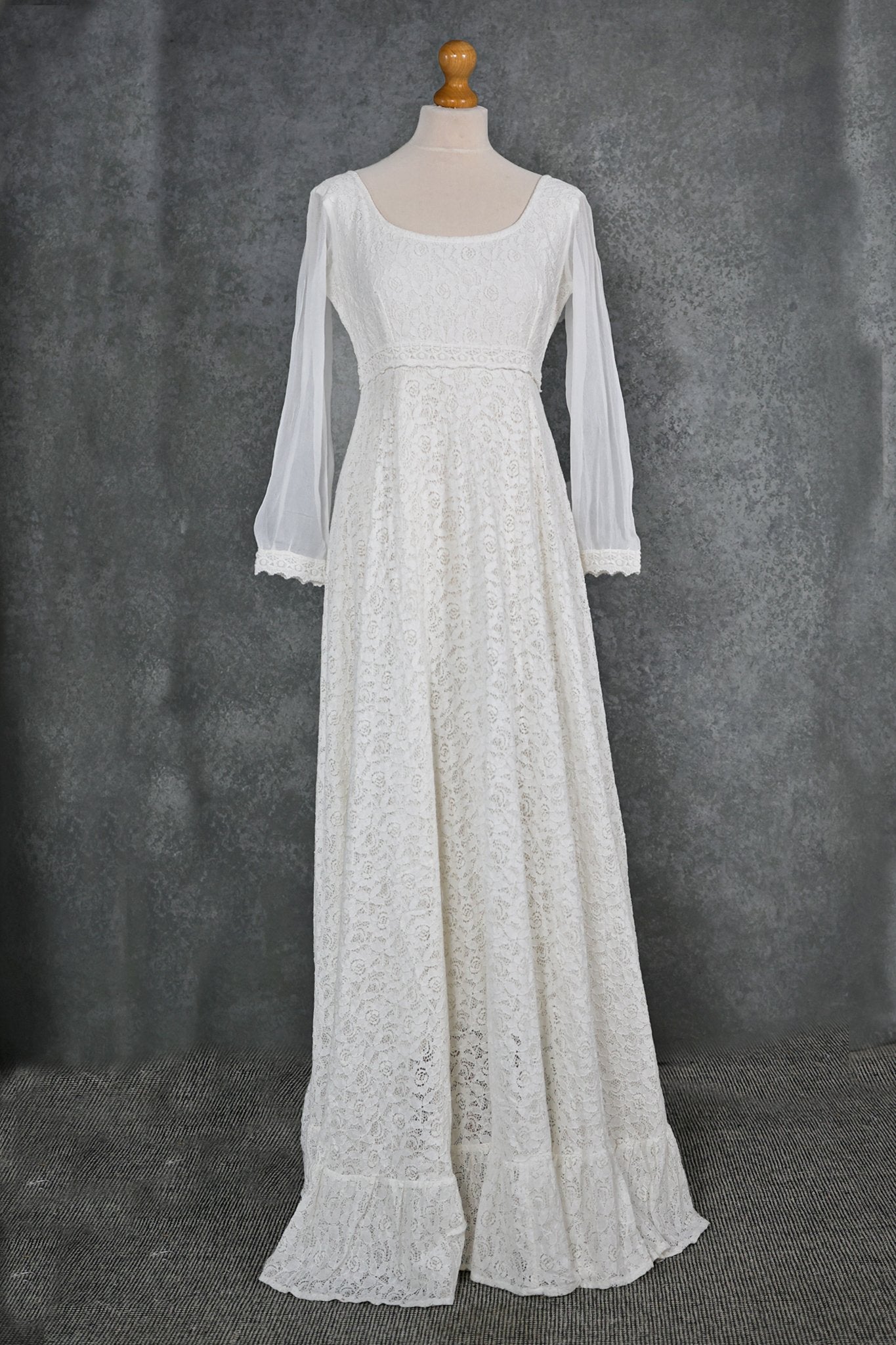 Ivory cotton lace sleeved wedding dress on mannequin with scoop neckline, empire waist detail and flowing hem frill