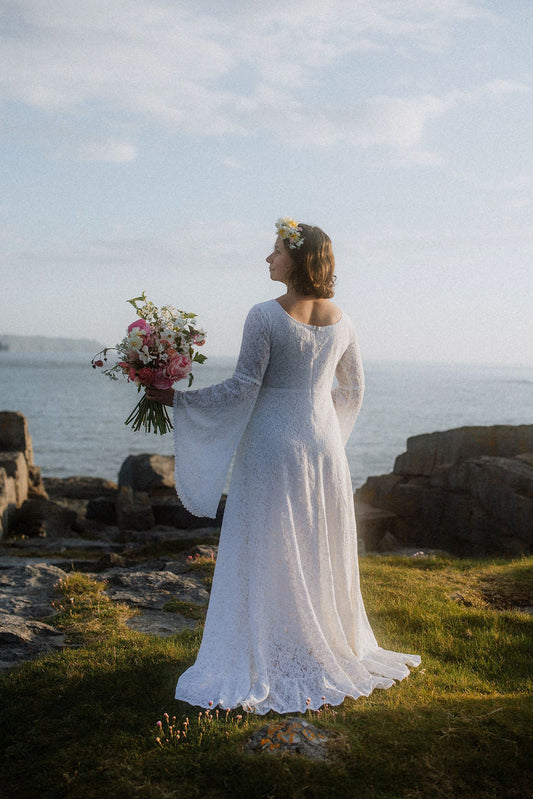 A woman in a white long-sleeve lace wedding gown stands outdoors by the sea, holding a pink and white bouquet, with rocks, grass, and a cloudy sky in the background.