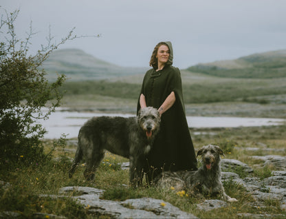 A woman in a long dark green felt wool cape stands outdoors on a rocky grassy landscape in the Irish burren with two large gray dogs, with hills and water in the background.