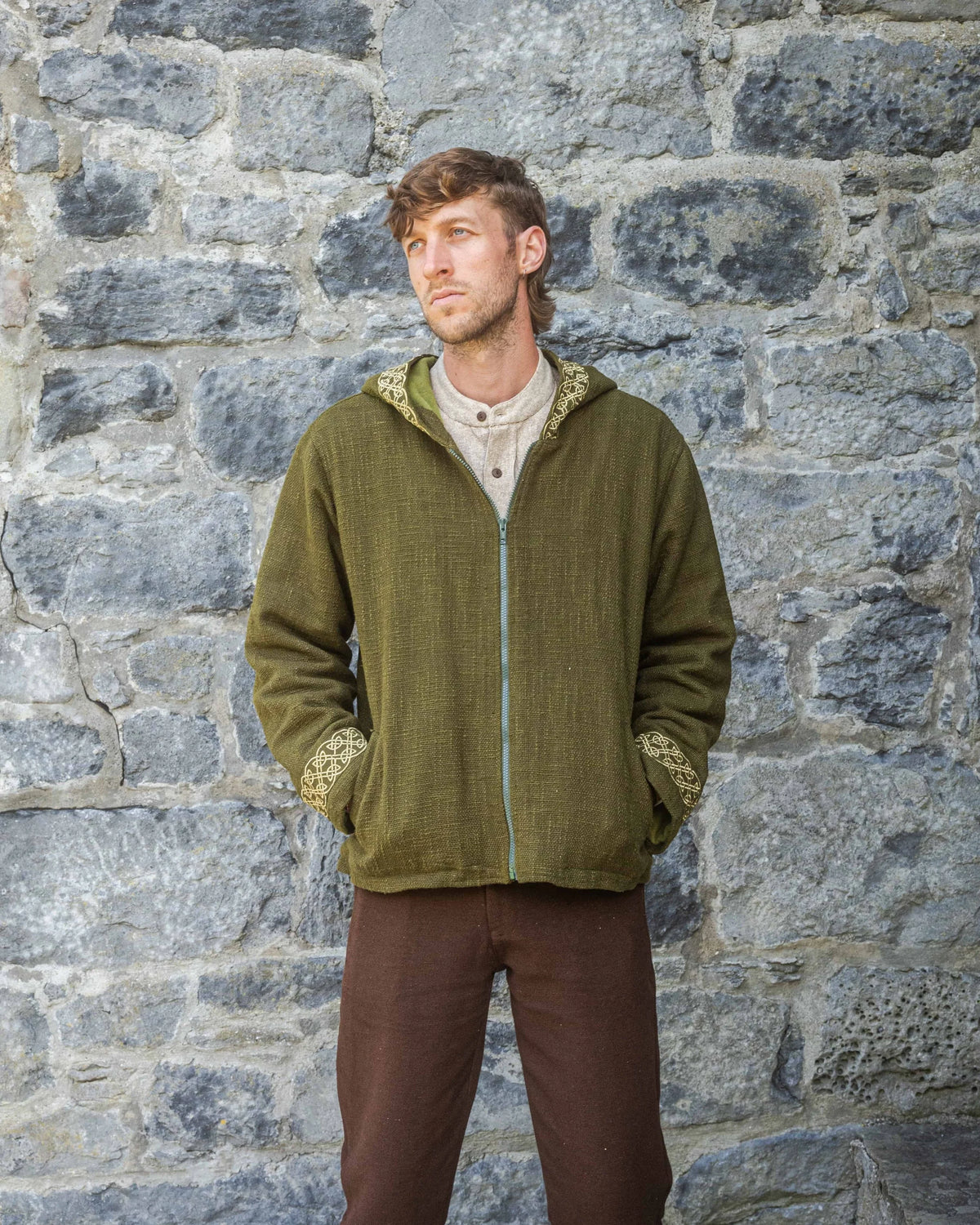 A man wearing the moss green Cernunnos Celtic Jacket stands against a stone wall. The jacket features a hood, golden Celtic knot embroidery on the cuffs and hood trim, and a front zip. Made in handwoven cotton with soft fleece lining for warmth.