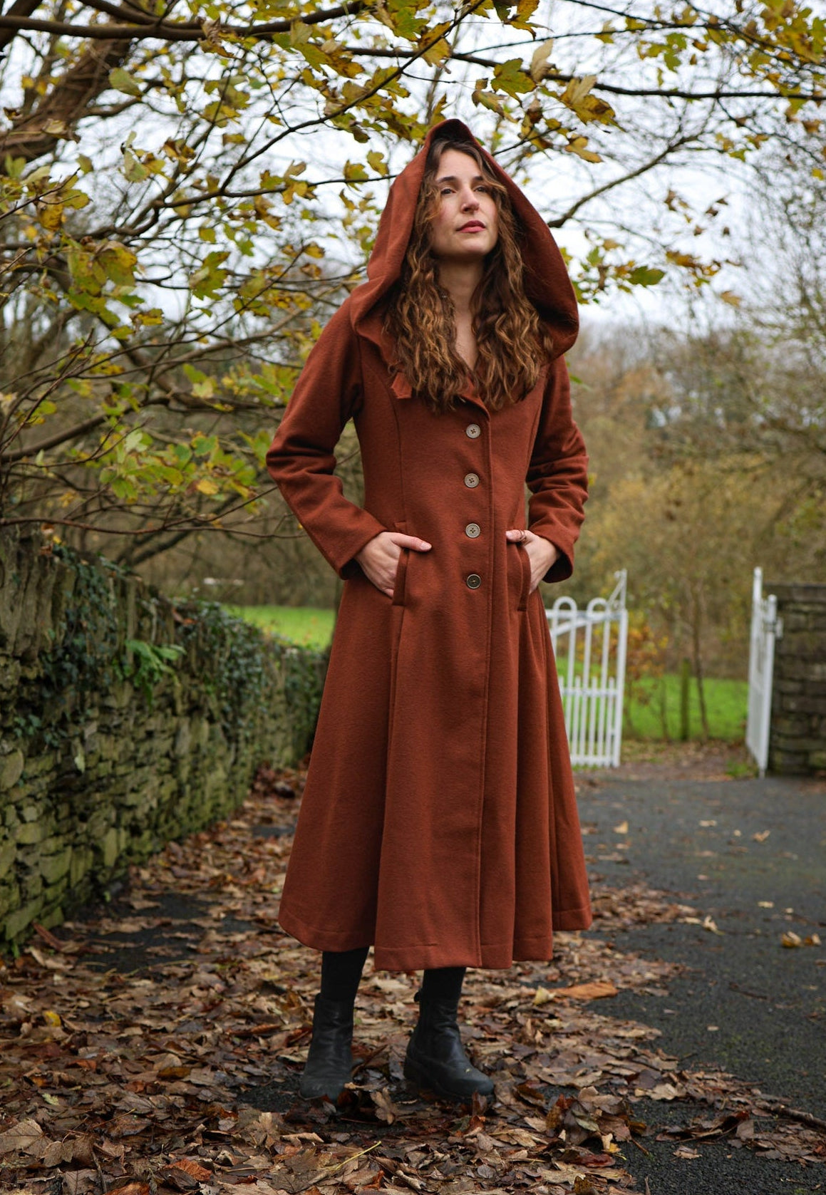 Woman wearing rusty red Princess maxi wool coat with large hood, pockets and full skirt in an autumn setting designed by Celtic Fusion Design