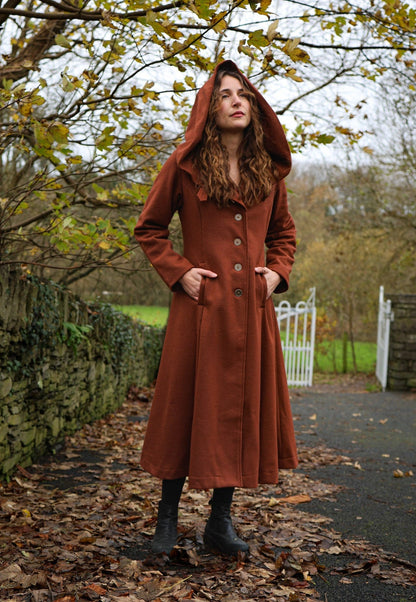 Woman wearing rusty red Princess maxi wool coat with large hood, pockets and full skirt in an autumn setting designed by Celtic Fusion Design