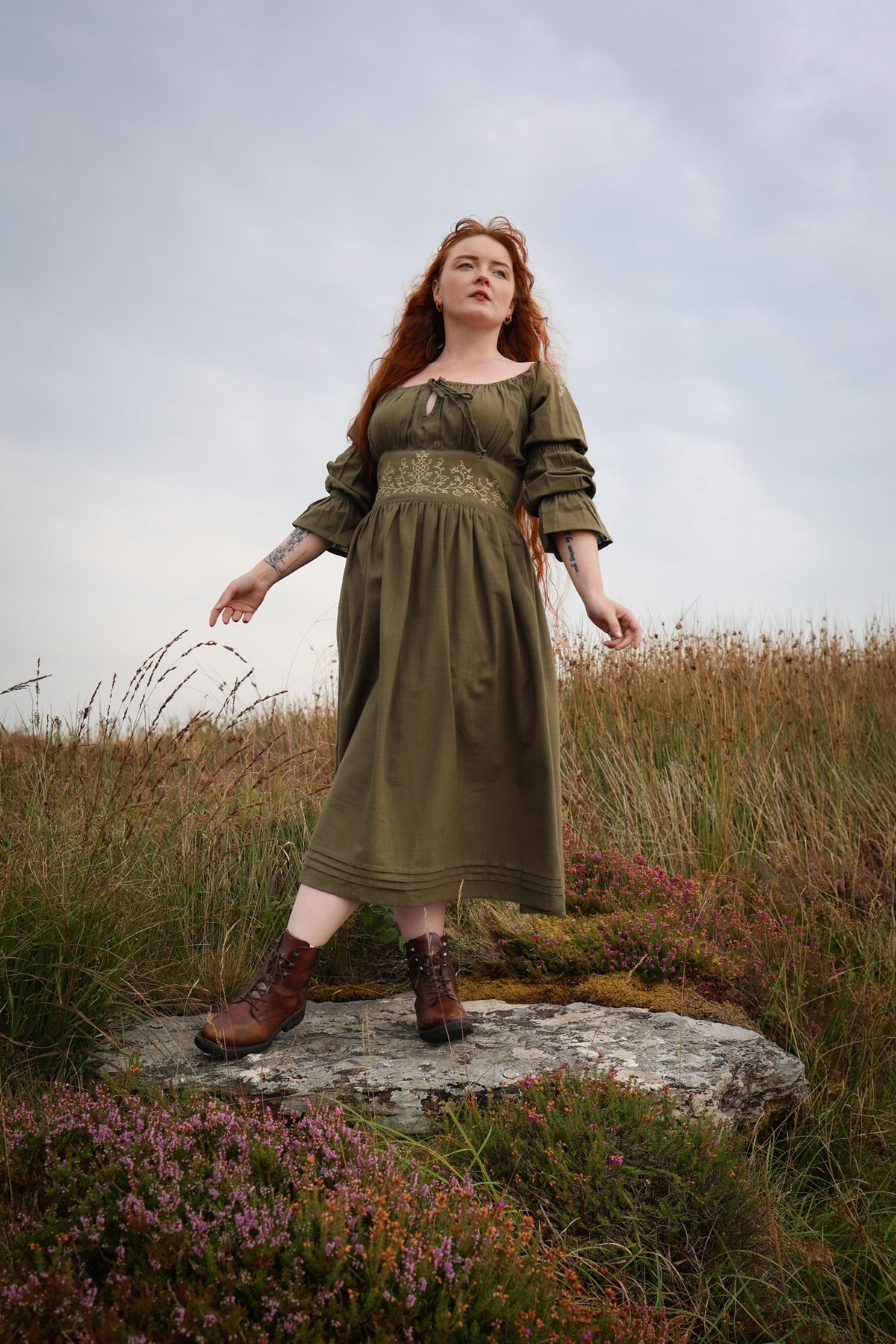Girl with red hair standing in a field of flowers wearing a sage green midi dress with long sleeves and floral embroidery waistband.