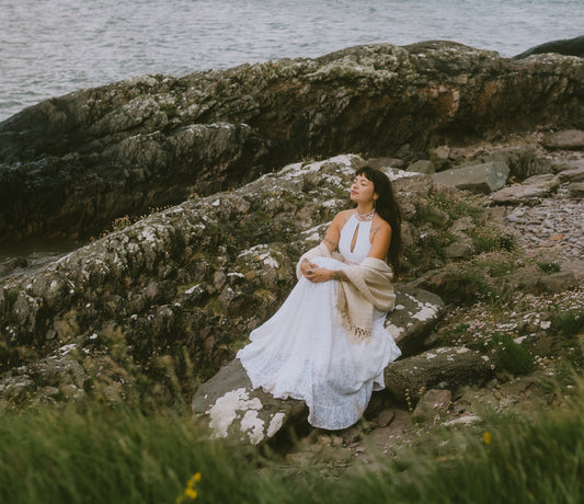 A woman in a white sleeveless natural wedding dress sits on a rocky shoreline by the water, holding a beige shawl draped over her lap.