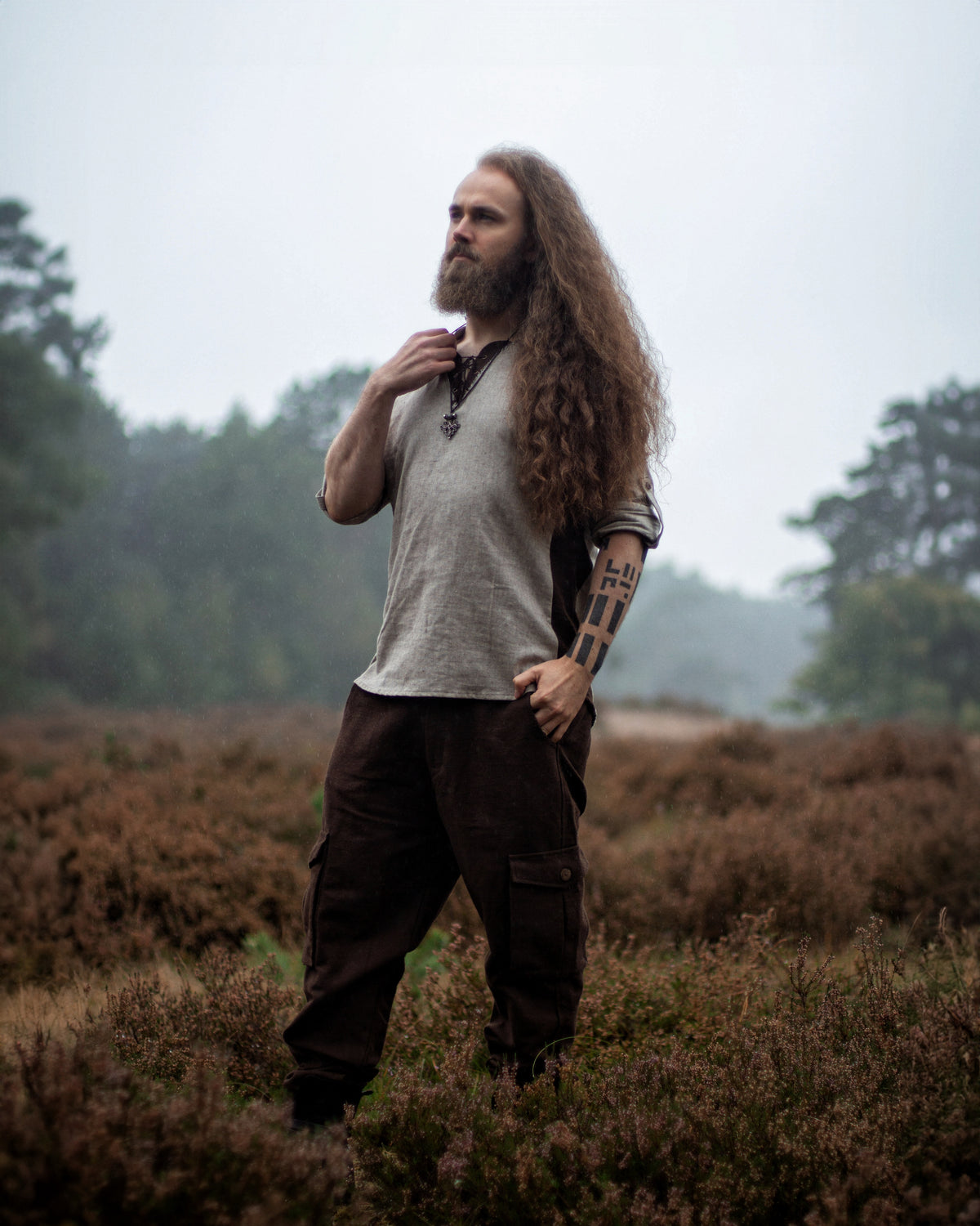 A man with long curly hair and a beard stands in a misty heather field, wearing a light gray shirt, dark brown cargo pants, and a black pendant necklace.